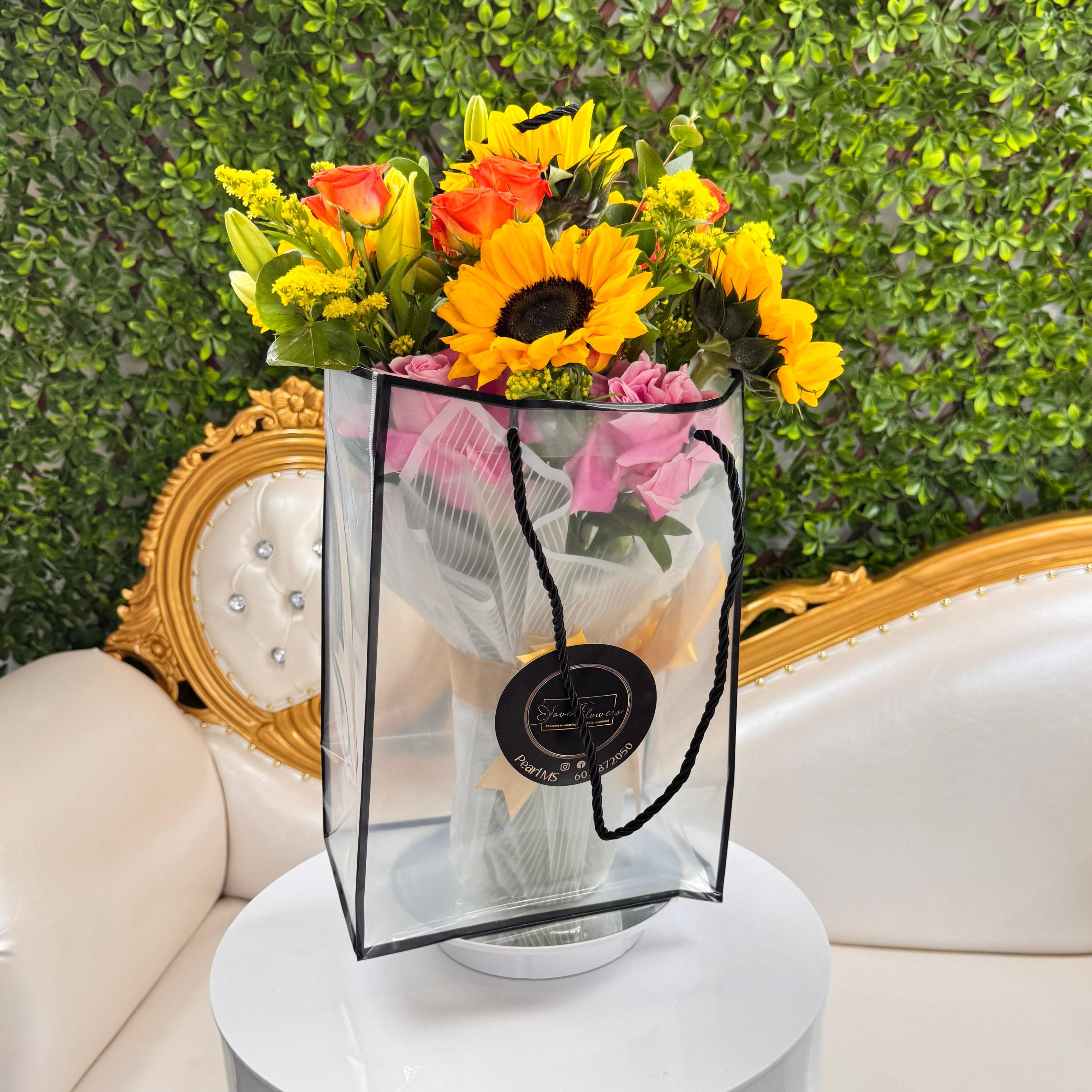 Bouquet of flowers in a clear bag on a white pedestal with a decorative chair and greenery in the background