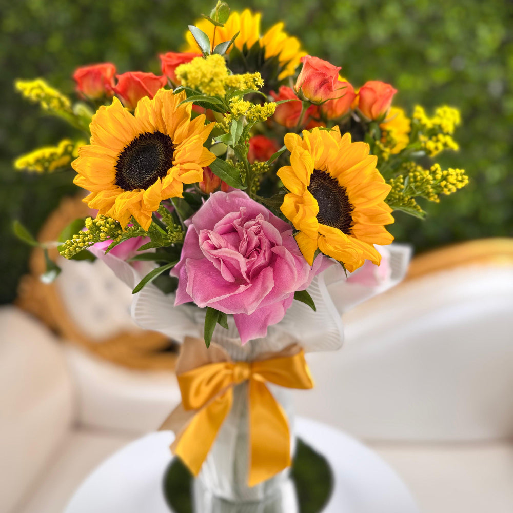 Bouquet of sunflowers and pink roses with a yellow ribbon in a white vase against a blurred green background.