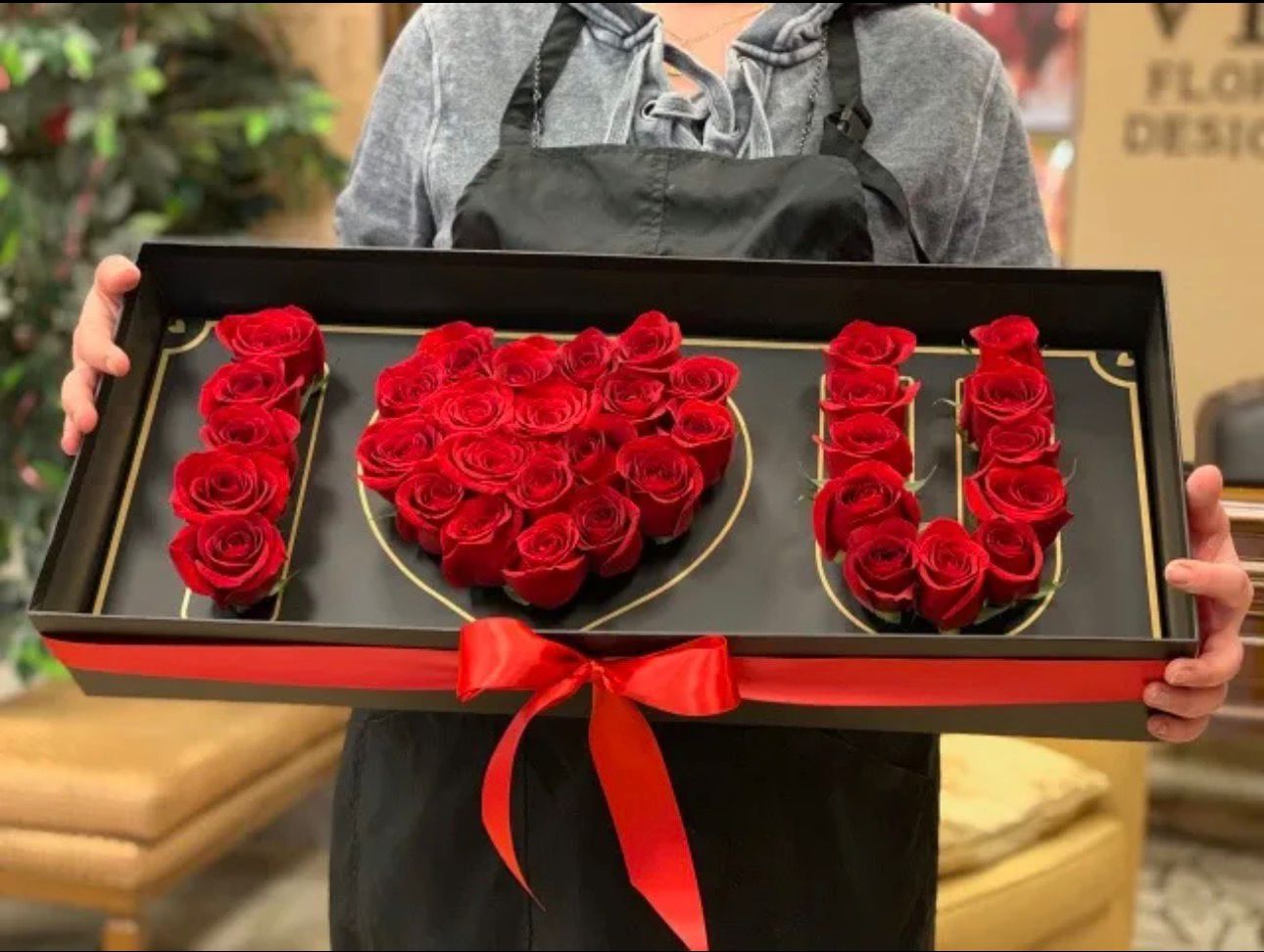 Person holding a decorative box with red roses shaped like a heart and letters spelling 'LOVE'.